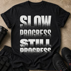 Black t-shirt with text "SLOW PROGRESS STILL PROGRESS" and gym accessories.