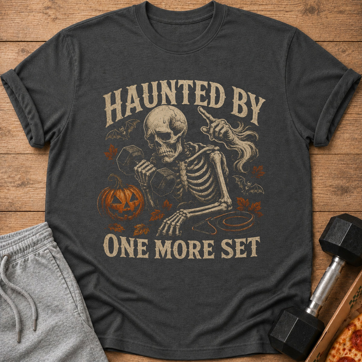 Dark grey t-shirt with skeleton lifting weights and "HAUNTED BY ONE MORE SET" text.