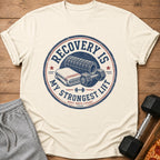 Cream t-shirt with "Recovery is My Strongest Lift" graphic and gym accessories.