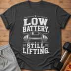 Dark gray t-shirt with "LOW BATTERY. STILL LIFTING." text and dumbbell graphic.