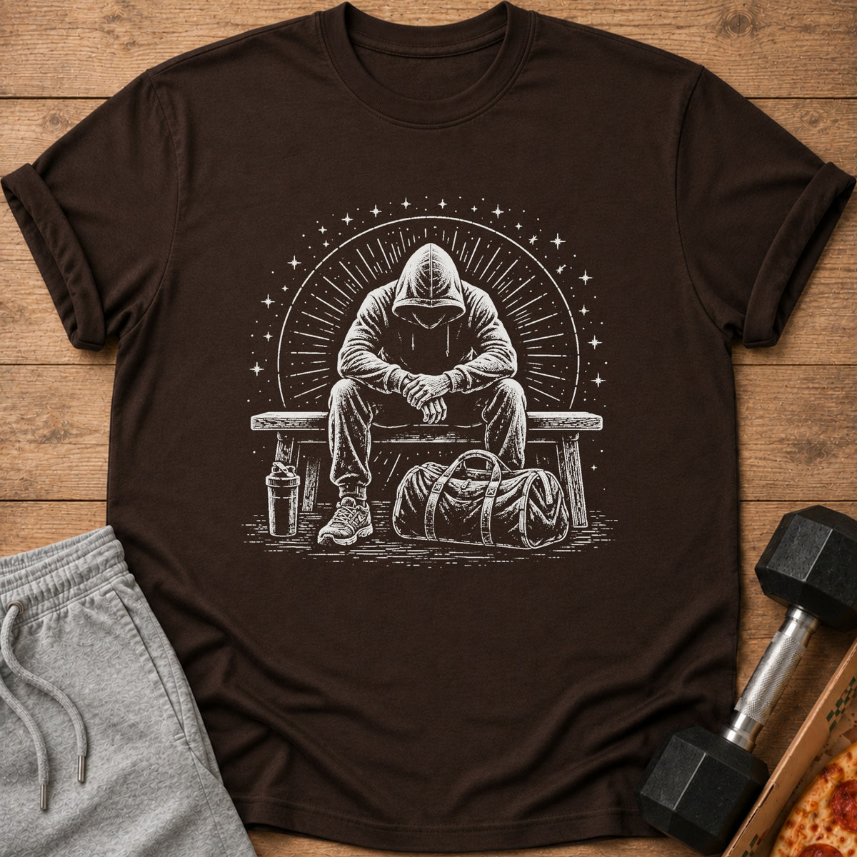 Brown t-shirt with a white graphic of a person in a hoodie sitting on a bench with gym bag.