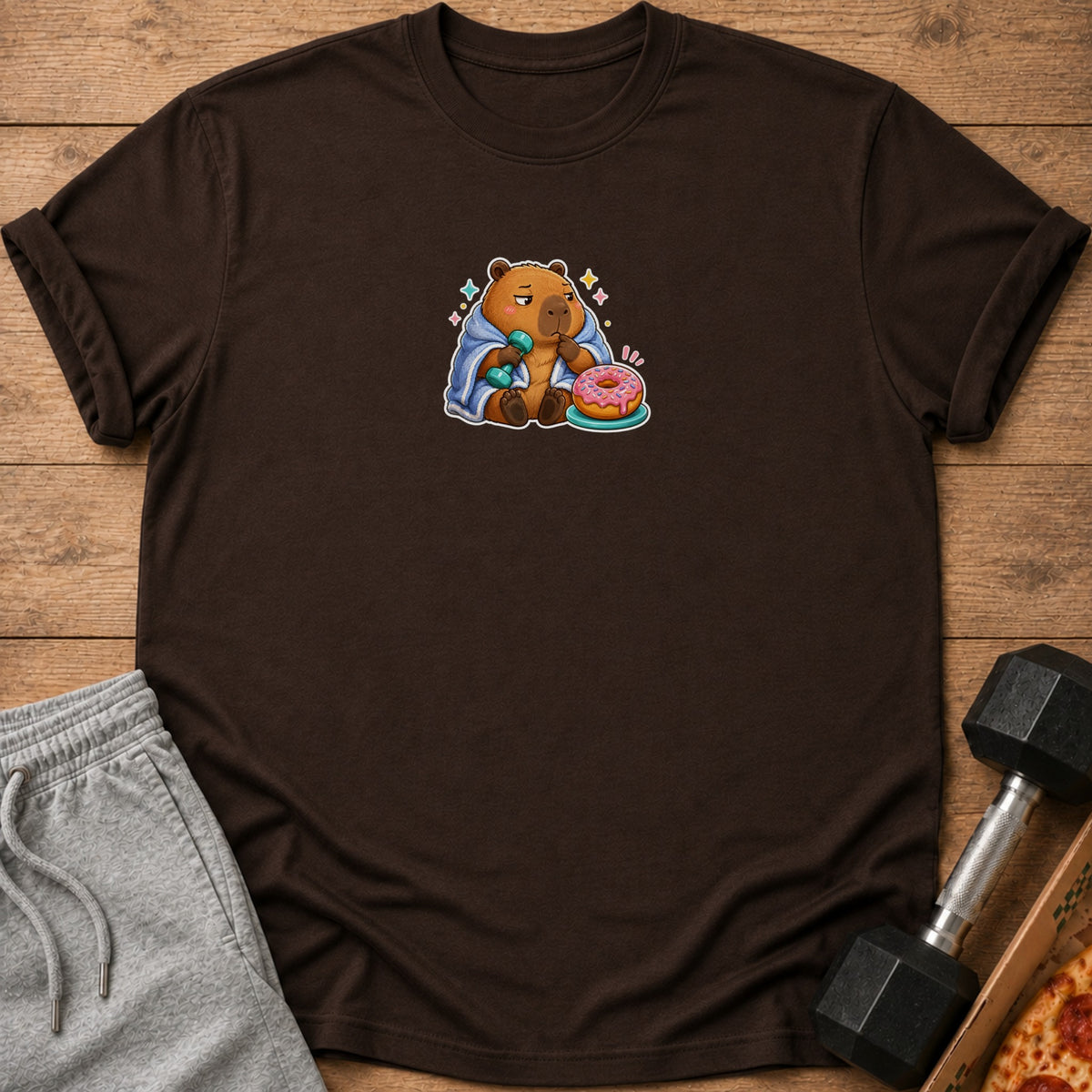 Brown t-shirt with a cartoon capybara wrapped in a blanket, holding a dumbbell and a donut.