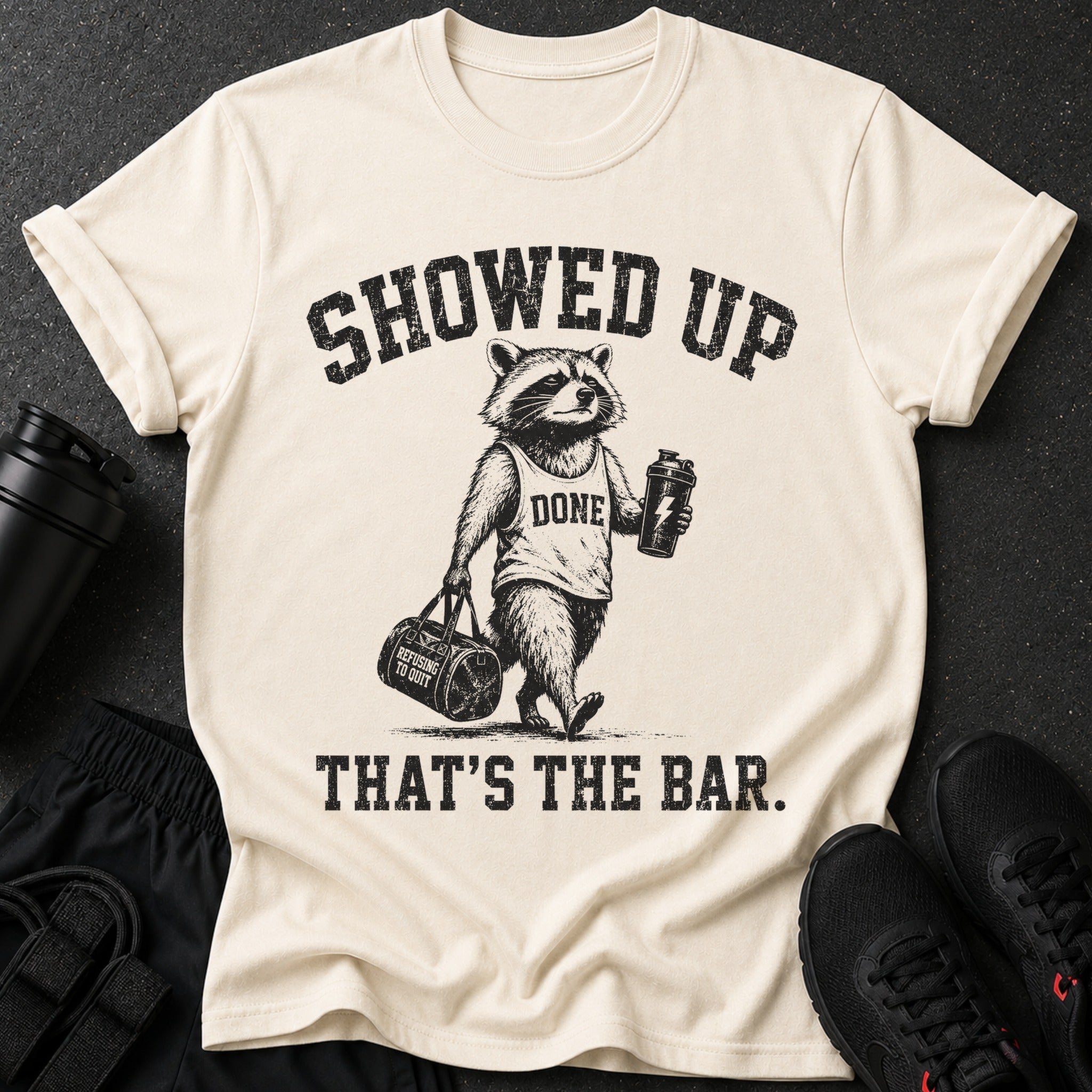 Graphic tee with raccoon holding gym bag and shaker. Text reads "SHOWED UP DONE THAT'S THE BAR."