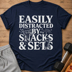 Navy t-shirt with white text and graphics reading "Easily Distracted By Snacks & Sets".