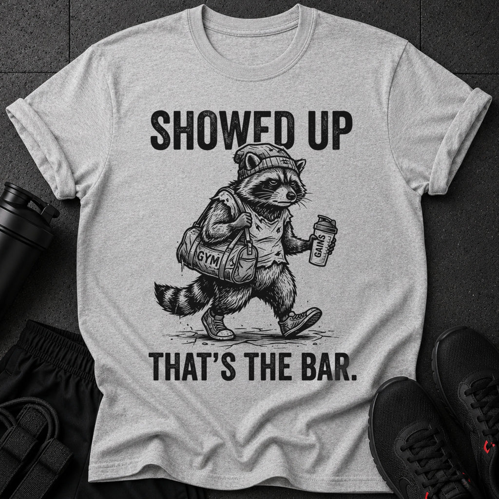 Graphic tee with a raccoon wearing a beanie, carrying a gym bag and shaker bottle. Text reads "SHOWED UP THAT'S THE BAR."