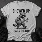 Graphic tee with a raccoon wearing a beanie, carrying a gym bag and shaker bottle. Text reads "SHOWED UP THAT'S THE BAR."