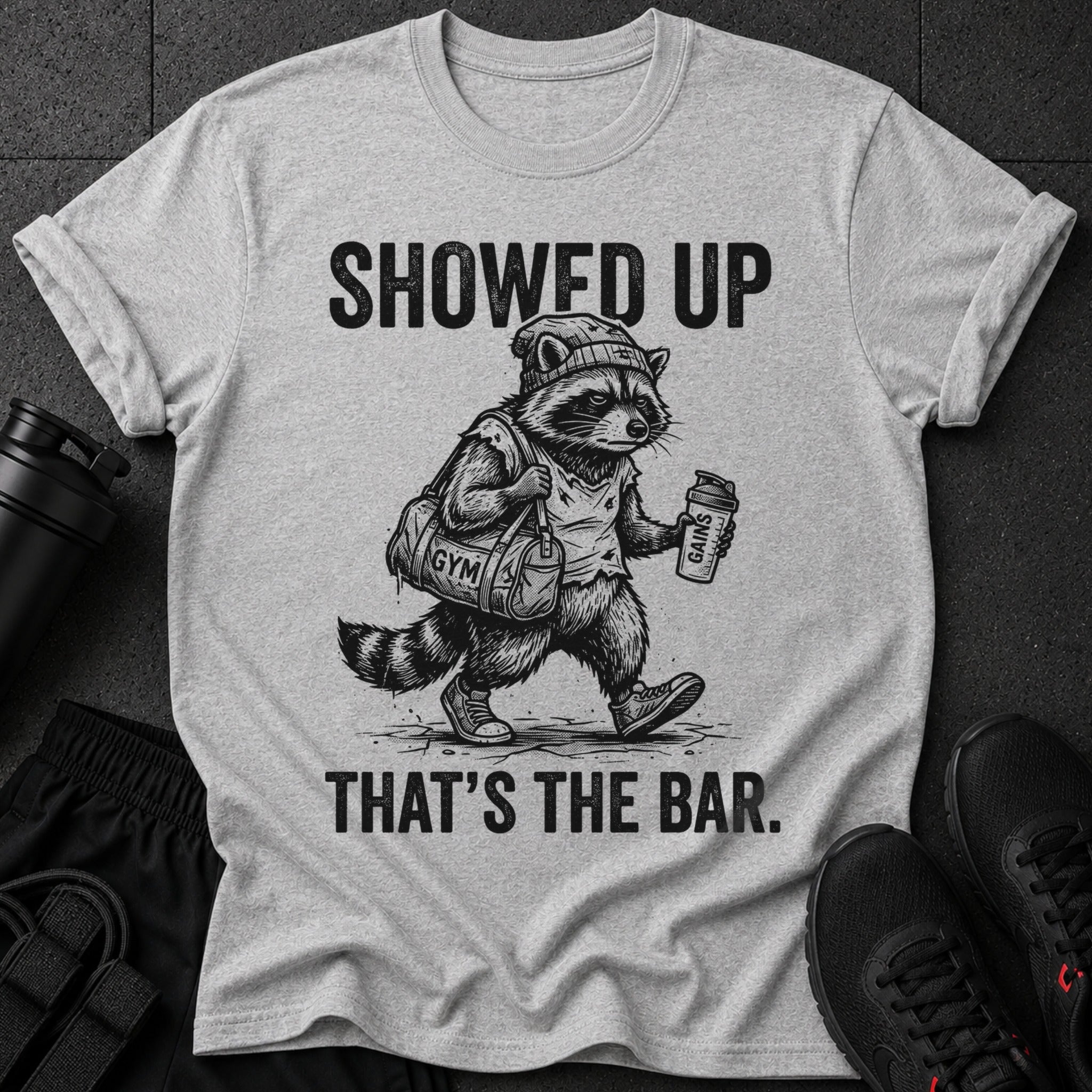 Graphic tee with a raccoon wearing a beanie, carrying a gym bag and shaker bottle. Text reads "SHOWED UP THAT'S THE BAR."