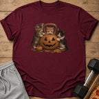 Three kittens interact with a jack-o'-lantern and kettlebell on a maroon t-shirt.