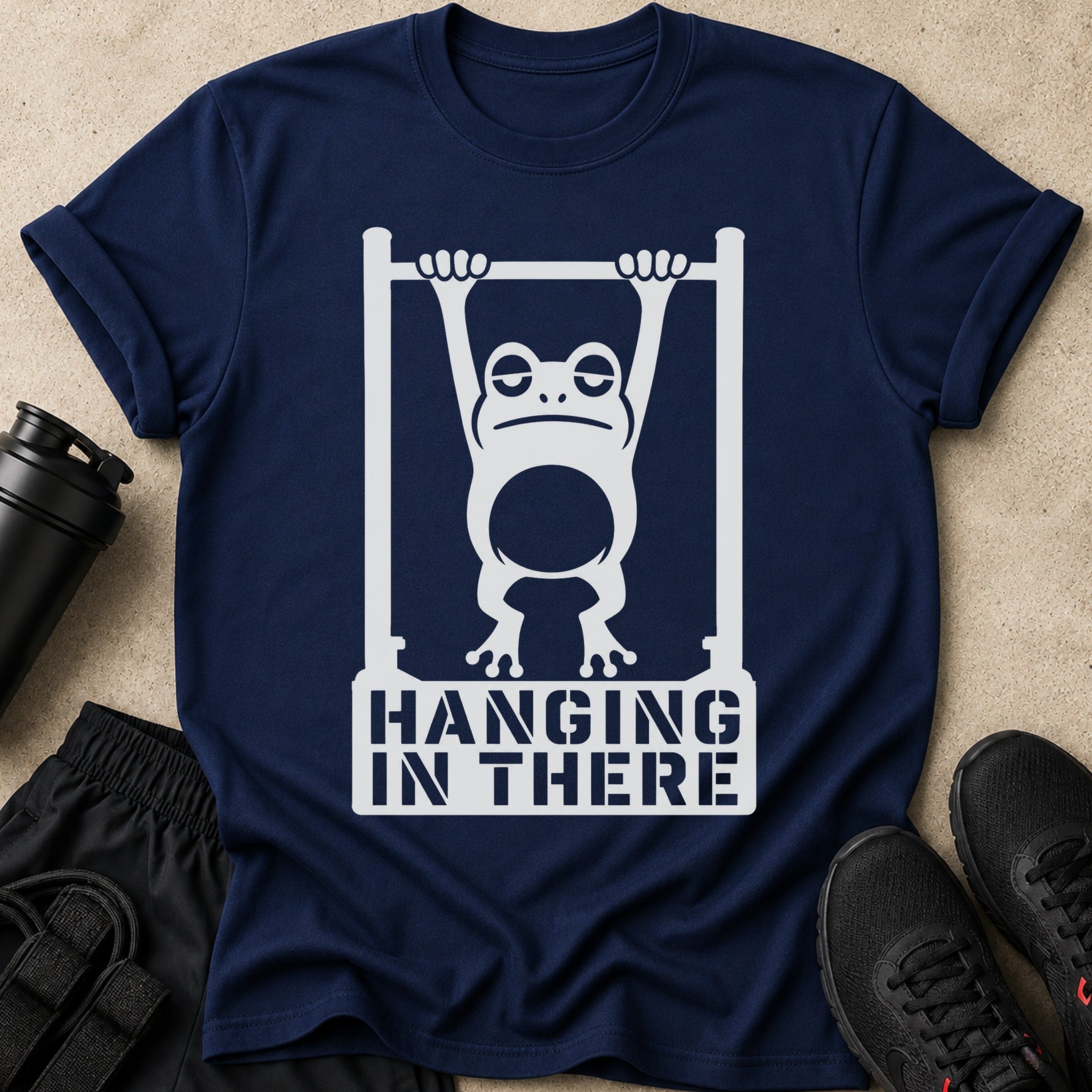 Navy t-shirt with white graphic of a tired frog hanging from a bar, text reads "HANGING IN THERE."
