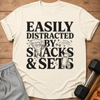 Cream t-shirt with black text "Easily Distracted By Snacks & Sets" and cartoon images.
