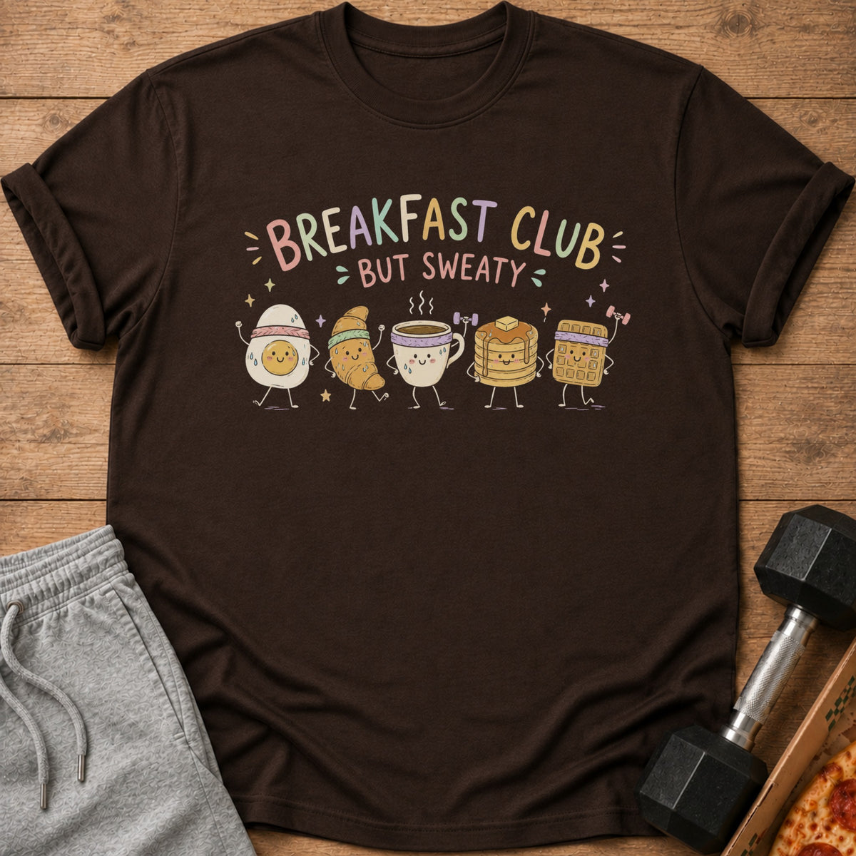 A brown t-shirt featuring a cartoon design of breakfast foods exercising with the text Breakfast Club But Sweaty.