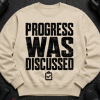 Beige sweatshirt with black text "PROGRESS WAS DISCUSSED" and a clipboard icon.
