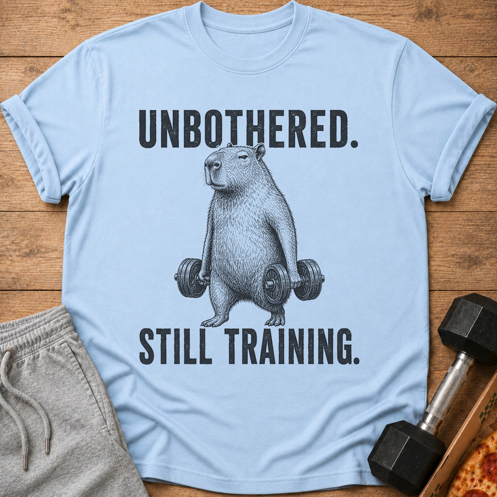 Light blue t-shirt with black text "UNBOTHERED. STILL TRAINING." and capybara lifting weights graphic.