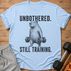 Light blue t-shirt with black text "UNBOTHERED. STILL TRAINING." and capybara lifting weights graphic.