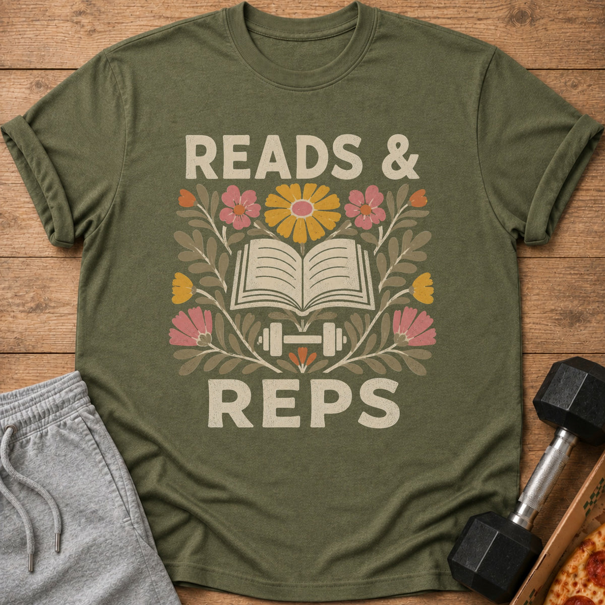 An olive green t-shirt with the text READS AND REPS surrounding an open book, dumbbell, and flowers graphic.