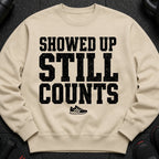 Light tan sweatshirt with black distressed text "SHOWED UP STILL COUNTS" and a sneaker graphic.