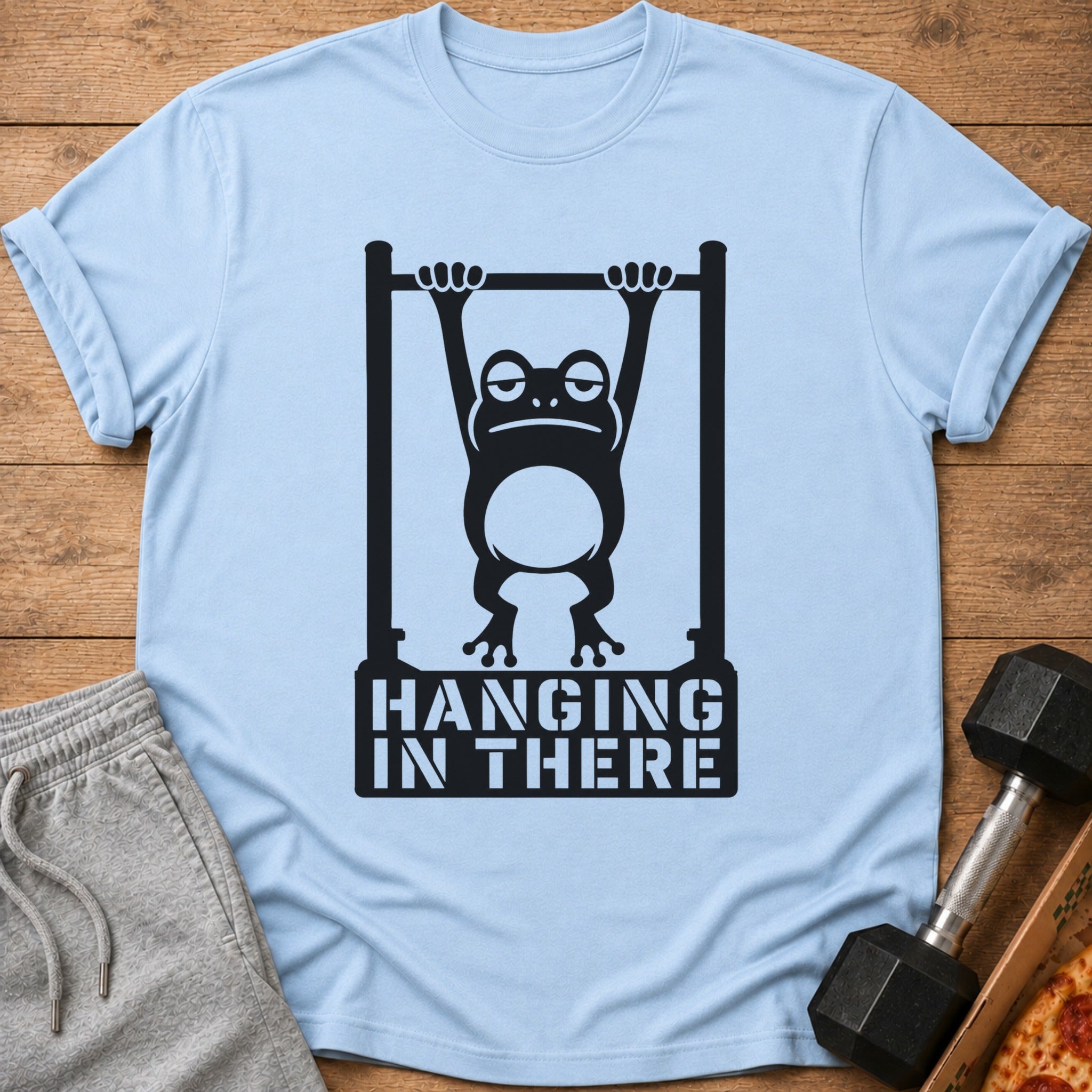 Light blue t-shirt with a black graphic of a tired frog hanging from a bar, text reads "HANGING IN THERE".