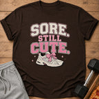 Brown t-shirt with text "SORE STILL CUTE" and a sneaker with a pink bow.