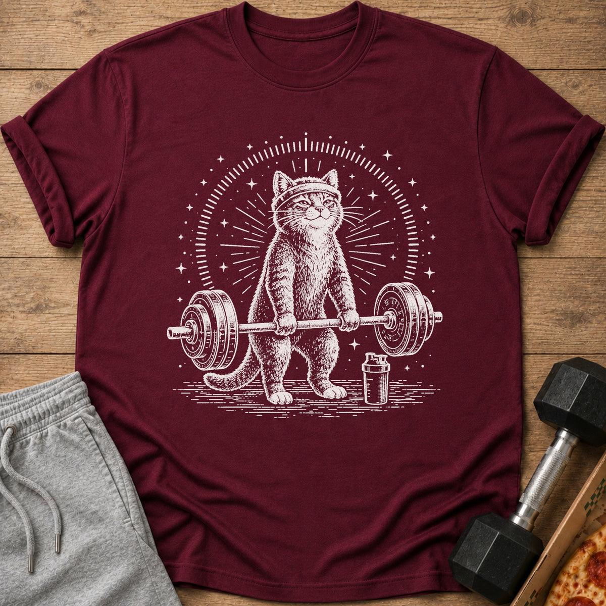 Maroon t-shirt with white drawing of a cat lifting a barbell, with stars and a shaker bottle.