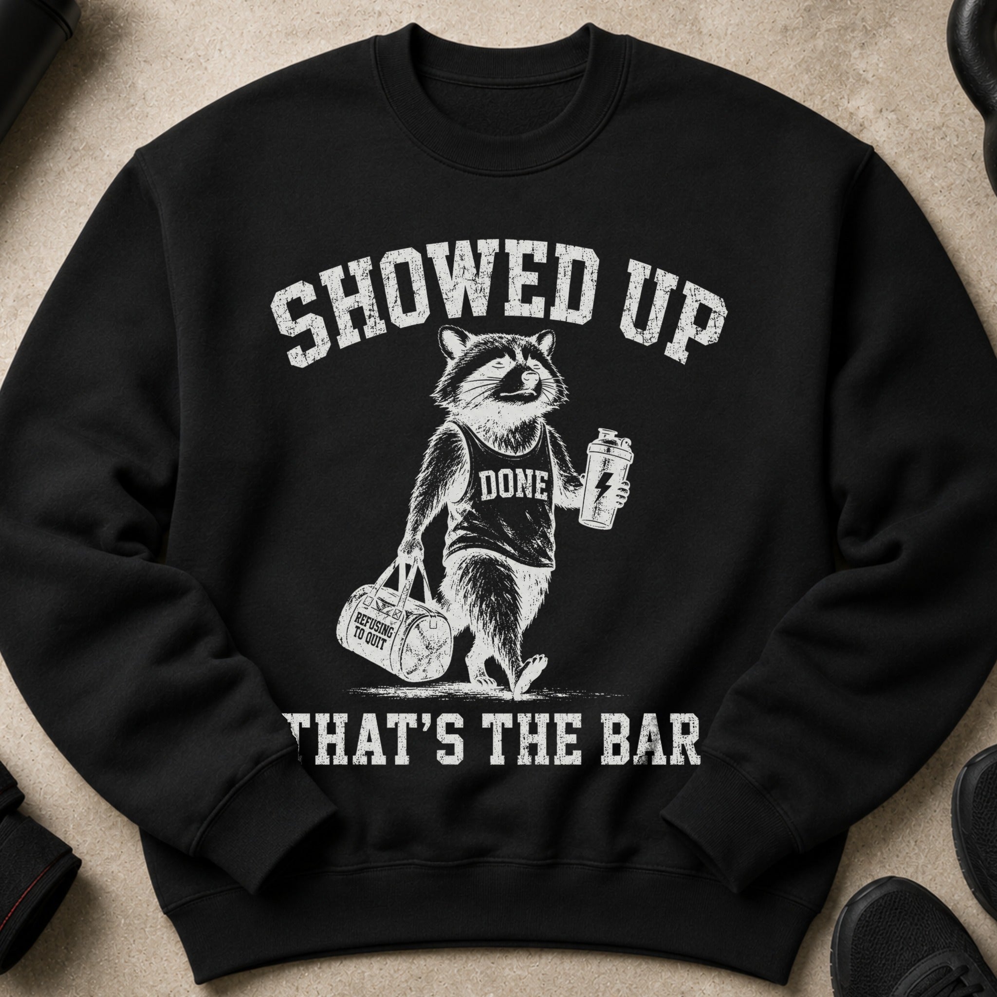 Black sweatshirt with distressed graphic of raccoon holding gym bag and shaker, reads "SHOWED UP...DONE...THAT'S THE BAR".