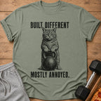 Green t-shirt with a cat holding a kettlebell and text "Built Different. Mostly Annoyed."