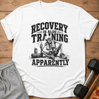 A white t-shirt featuring a black graphic of a skeleton giving a thumbs-up next to workout gear, with the text RECOVERY IS ALSO TRAINING APPARENTLY.