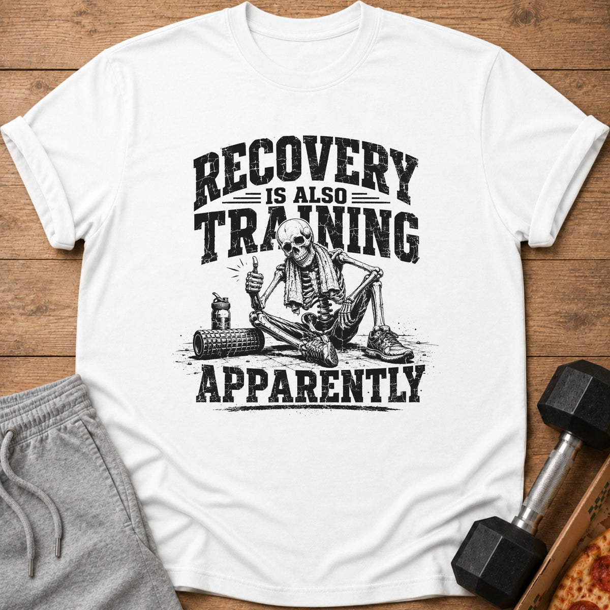 A white t-shirt featuring a black graphic of a skeleton giving a thumbs-up next to workout gear, with the text RECOVERY IS ALSO TRAINING APPARENTLY.