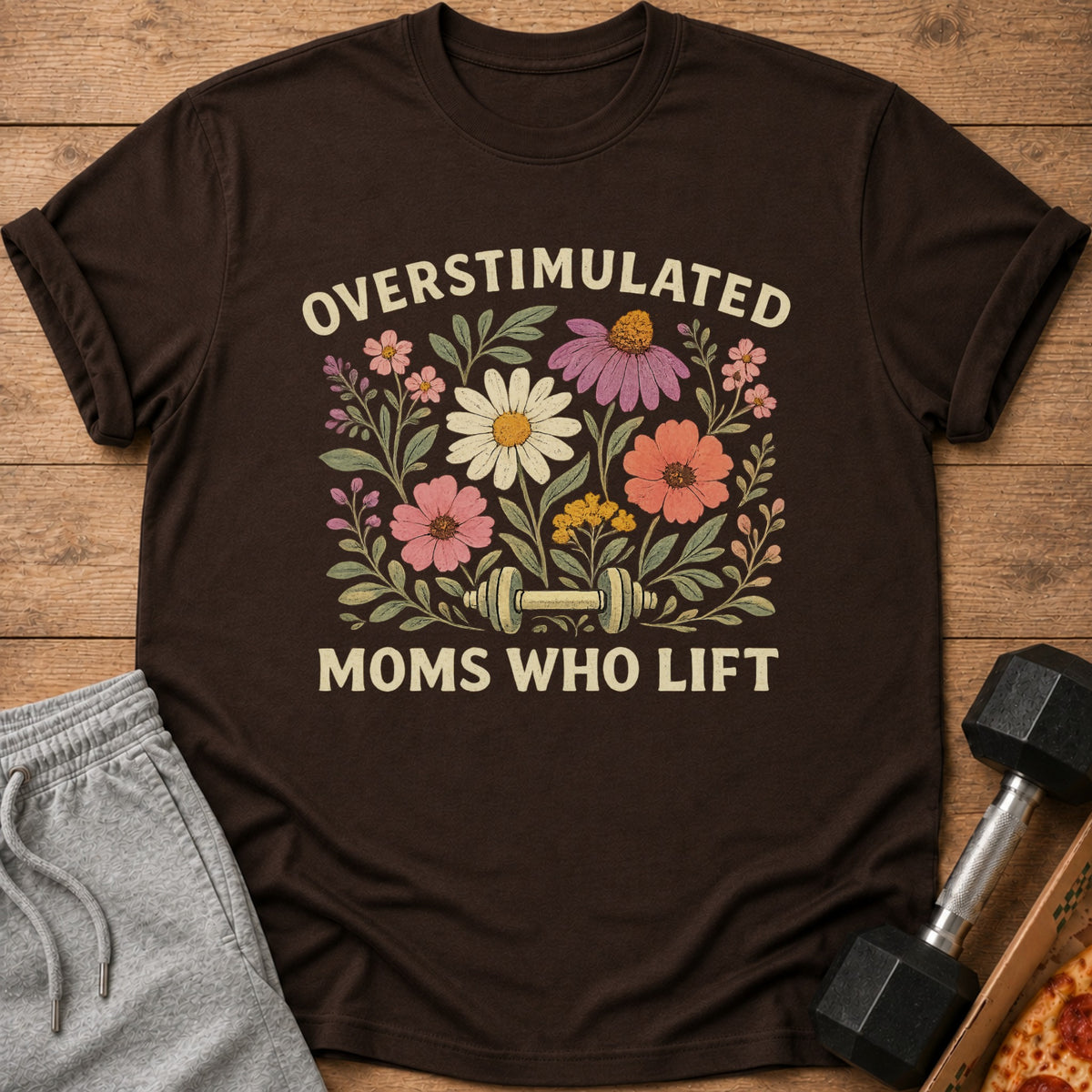 A brown t-shirt featuring floral graphics, a dumbbell, and the text Overstimulated Moms Who Lift.