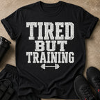 Black t-shirt with white distressed text reading "TIRED BUT TRAINING" and a dumbbell graphic.