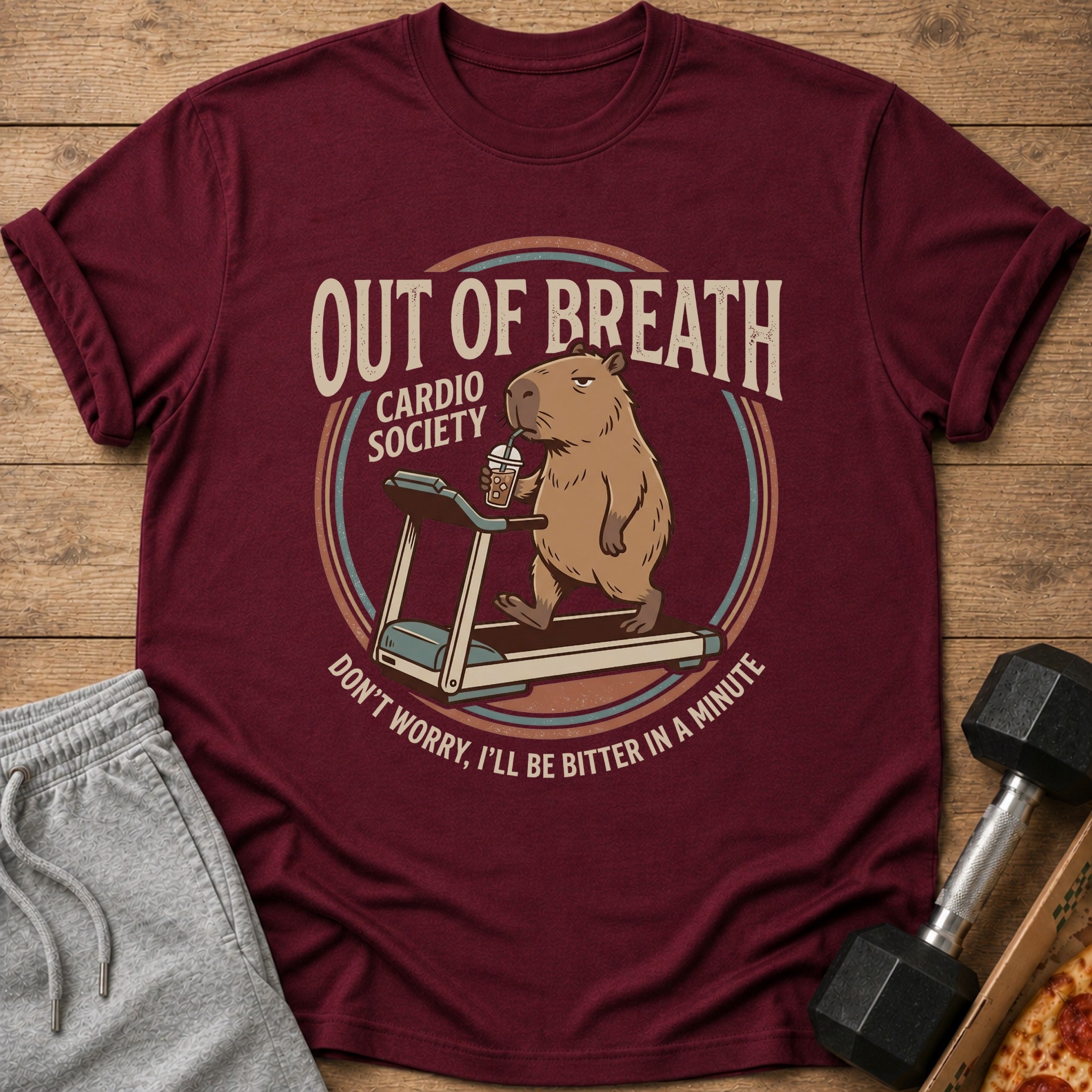 Maroon t-shirt graphic of a capybara on a treadmill, drinking a beverage.
