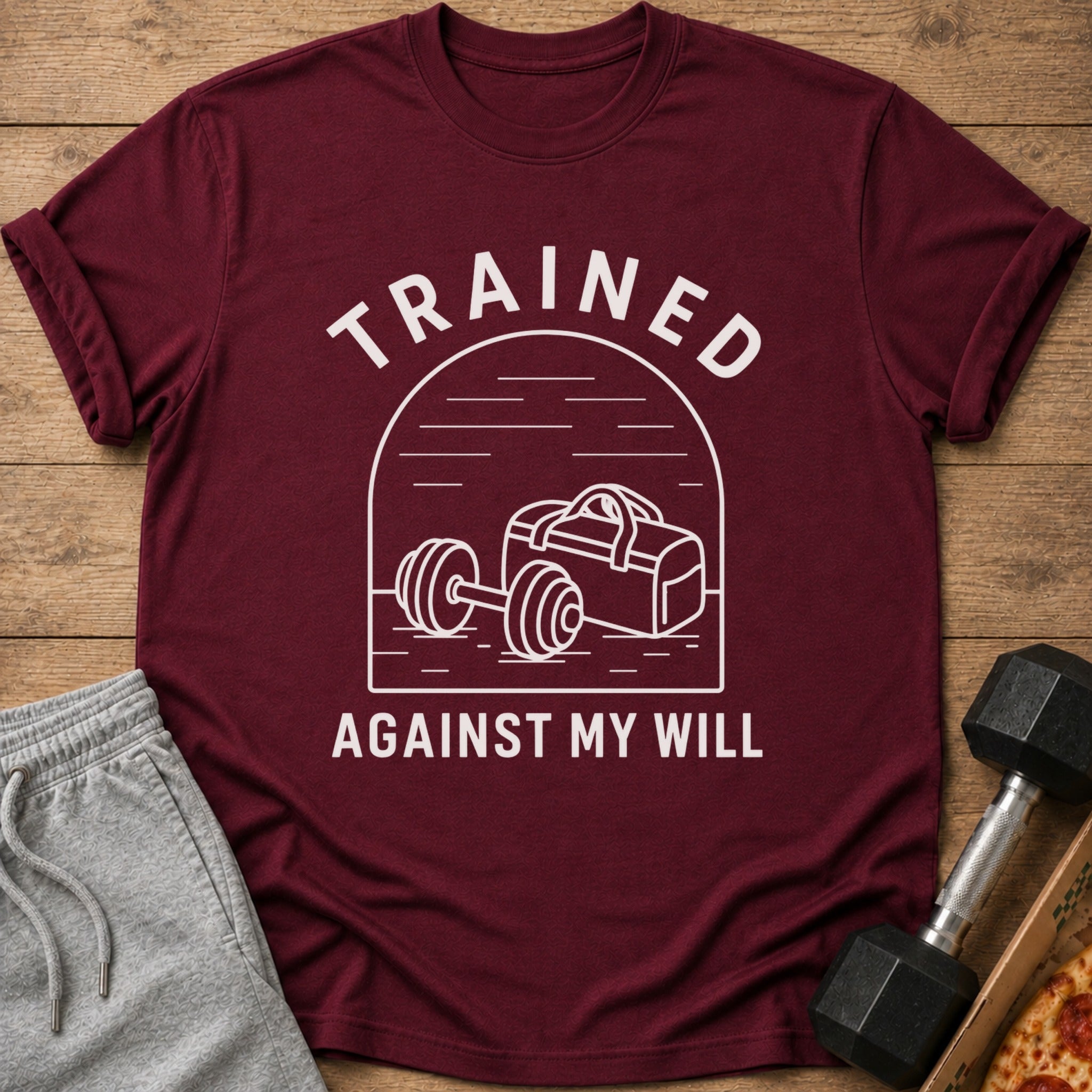 Maroon t-shirt with white text "TRAINED AGAINST MY WILL" and gym graphic.