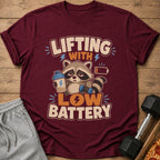 T-shirt with cartoon raccoon holding shaker and dumbbell, text "Lifting With Low Battery".
