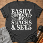 Dark gray t-shirt with white text "Easily Distracted By Snacks & Sets" and cartoon graphics.