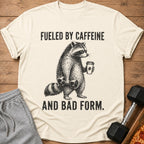 T-shirt graphic of a raccoon holding a dumbbell and coffee cup, text "Fueled by Caffeine and Bad Form."