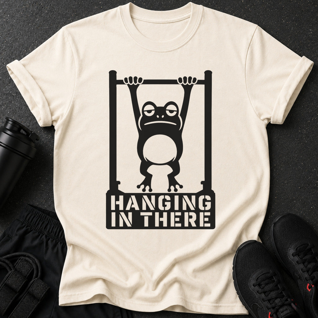 Cream t-shirt with a black graphic of a frog hanging from a bar, text reads "HANGING IN THERE".