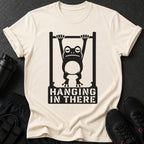 Cream t-shirt with a black graphic of a frog hanging from a bar, text reads "HANGING IN THERE".