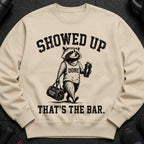 Light tan crewneck sweatshirt with a graphic of a raccoon holding a shaker bottle and gym bag.