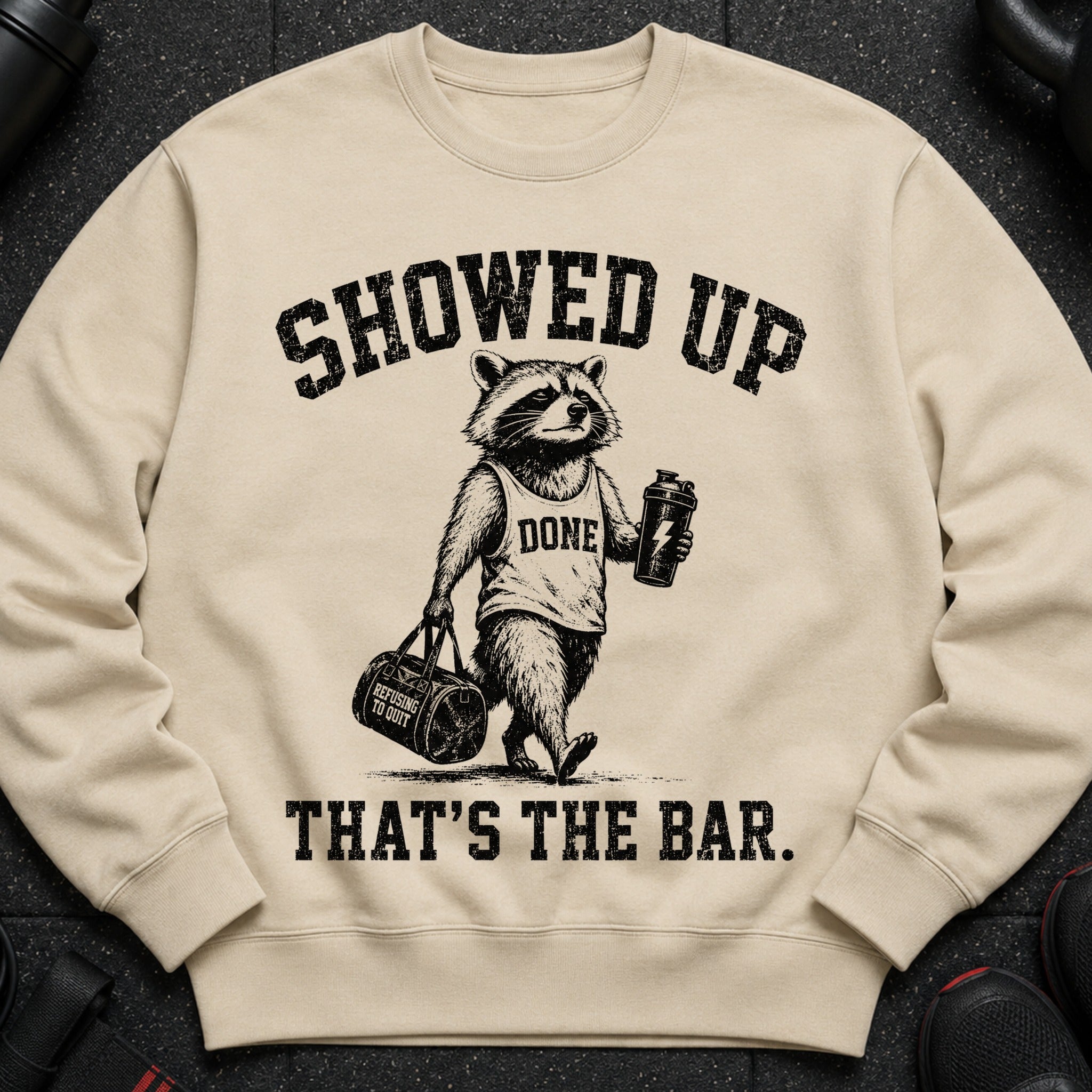 Light tan crewneck sweatshirt with a graphic of a raccoon holding a shaker bottle and gym bag.