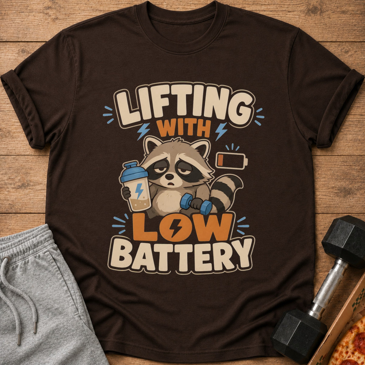 T-shirt graphic of raccoon with dumbbell and protein shake, text reads "Lifting with Low Battery".