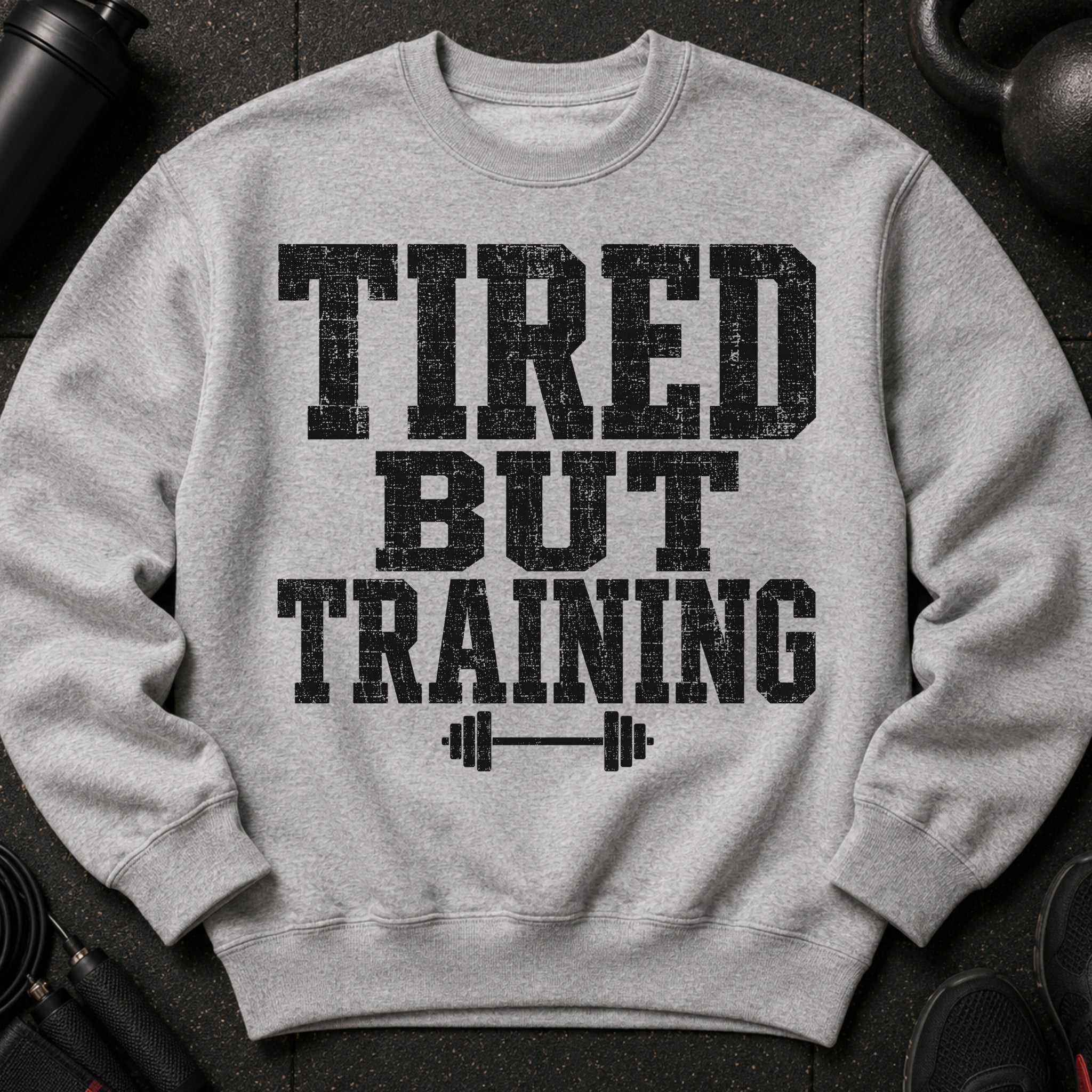 Gray sweatshirt with black distressed text "TIRED BUT TRAINING" and barbell graphic.