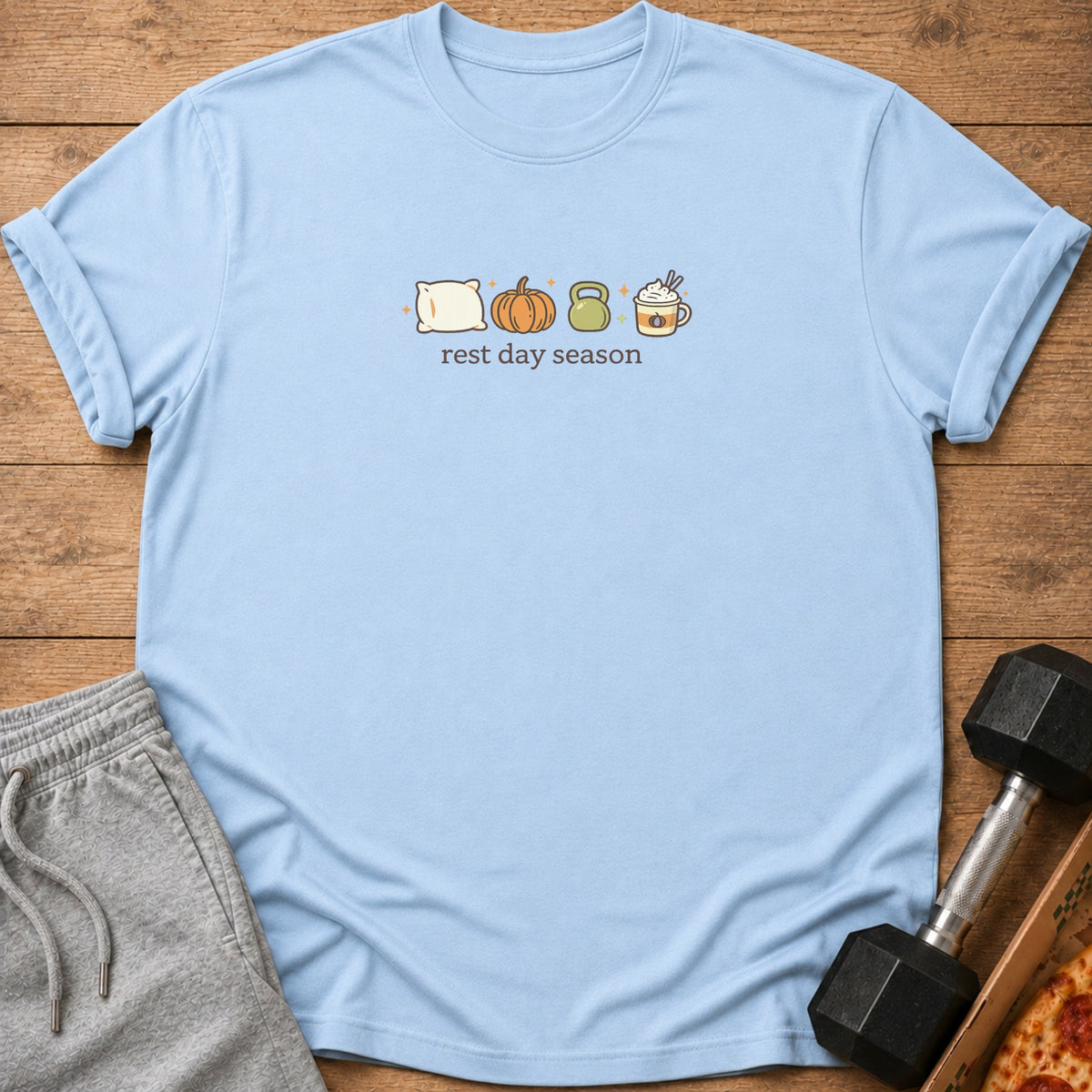 Light blue t-shirt with a graphic of a pillow, pumpkin, kettlebell, and coffee cup above "rest day season" text.