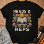 A black t-shirt printed with the text Reads & Reps, an open book, a dumbbell, and colorful flowers.