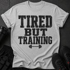Grey t-shirt with black distressed text "TIRED BUT TRAINING" and barbell graphic.