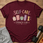A maroon t-shirt with a "Self-Care Strength Club" graphic, paired with gray shorts, a dumbbell, and pizza on a wooden background.