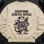 Light tan crewneck sweatshirt with a cartoon possum tangled in purple rope and the text "OVERTHINK. STRETCH. REPEAT."