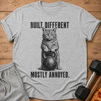 Gray t-shirt with a grumpy cat holding a kettlebell, text reads "BUILT DIFFERENT. MOSTLY ANNOYED."