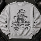 Grey sweatshirt with black graphic of a possum struggling with resistance bands, text "Stretched Thin, Still Here".