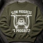 Green sweatshirt with white graphic of sloth hanging from bar, text reads "Slow Progress is Progress."