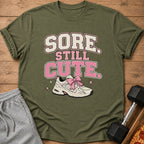 Olive green t-shirt with "SORE STILL CUTE" text and a sneaker with a pink bow.