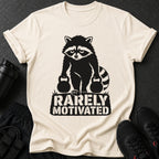 Cream t-shirt with a black graphic of a raccoon lifting two kettlebells, text reads "RARELY MOTIVATED."